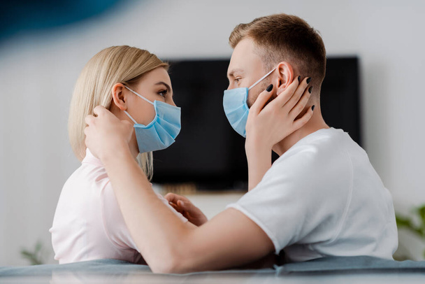 couple in medical masks looking at each other in living room  - Фото, изображение