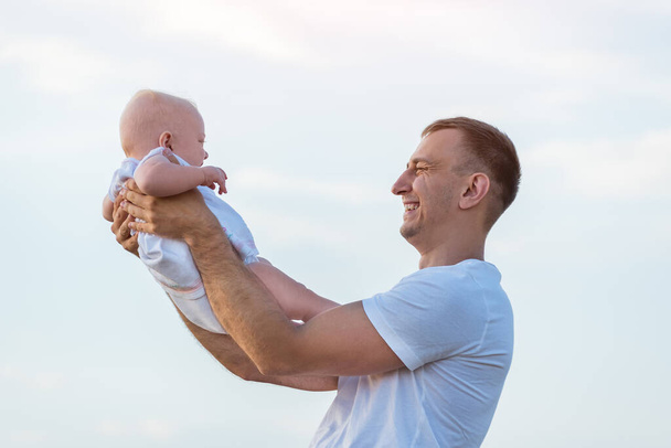 Ευτυχισμένος νεαρός πατέρας που κρατάει το μωρό στον ουρανό. Πορτρέτο του μπαμπά και του παιδιού - Φωτογραφία, εικόνα