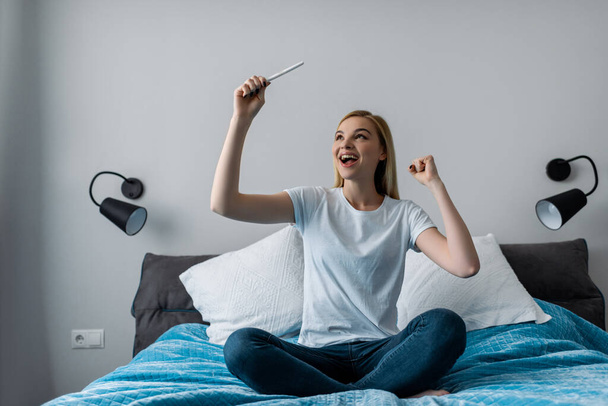 excited woman holding pregnancy test with positive result and sitting on bed - Valokuva, kuva