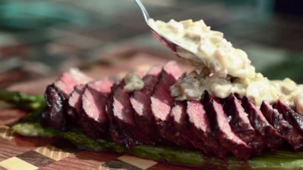Delicious sliced beef tenderloin with steamed asparagus and tasty mushrooms sauce on wooden board. Man pouring creamy condiment sauce with spoon on medium rare roasted beef steak, chef preparing - Footage, Video