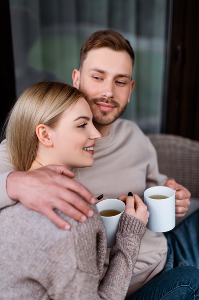selective focus of happy man holding cup of tea and hugging girlfriend  - 写真・画像