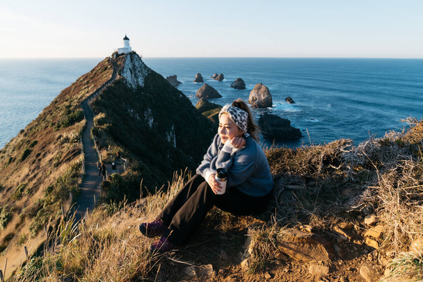 Krásný asijský turista odpočívající na Nugget Point, Dunedin, Nový Zéland. Mladý asijský cestovatel má rád kávu v dopoledních hodinách na pobřeží Nového Zélandu. Životní styl obrazu lidí v přírodní krajině. - Fotografie, Obrázek