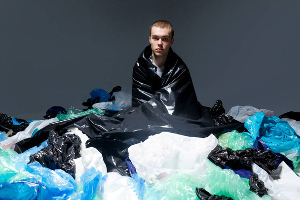 Atrakcyjny młody człowiek z fryzurą siedzącą i pokrytą plastikowym workiem śmieci, ciemne światło tła. - Zdjęcie, obraz
