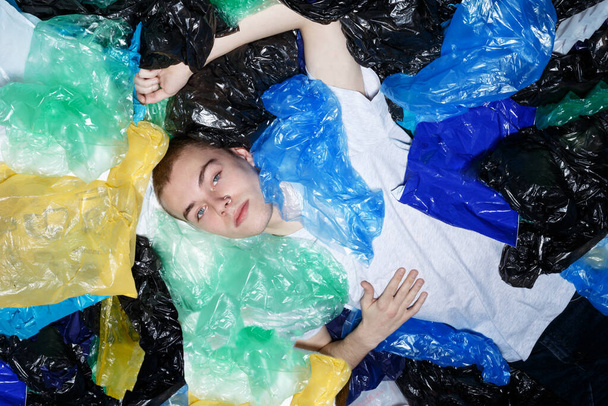 A hipster guy with blue eyes, lying down, with ring in nose, surrounded by plastic bags, looking up. Horizontal view. - Photo, Image