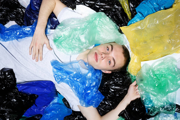 Young man with blue eyes, lying down, with ring in nose, surrounded by plastic bags, looking at camera. - Фото, изображение