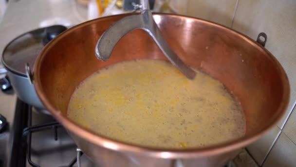 Preparation of traditional winter food at home, italian cuisine and ancient rustic recipe. Man pouring corn flour in cooper cauldron with boiling water and automatic metal paddle stirring yellow - Footage, Video
