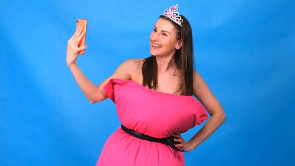 The most beautiful girl in a pink dress from pillows makes a selfie on a blue background. Crazy quarantine. Fashion 2020. Put on a pillow. Challenge 2020 due to house isolation - Фото, зображення