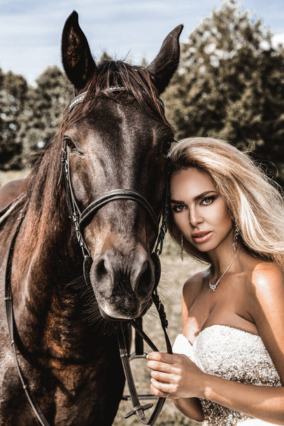 暗い馬と女性の肖像画。馬と美しい魅力的な女性。馬と結婚式のドレスで美しいブロンドの花嫁の肖像. - 写真・画像