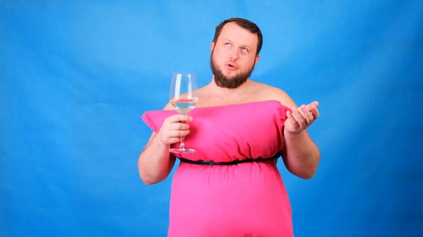 Homme barbu drôle dans une robe rose faite d'oreillers boit du vin d'un verre sur un fond bleu. Quarantaine folle. Mode 2020. Mets un oreiller. Défi 2020 dû à l'isolement des maisons
 - Photo, image