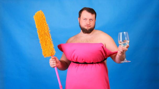 Homme barbu drôle dans une robe d'oreiller rose avec une serpillière et un verre de vin sur un fond bleu. Quarantaine folle. Drôle de ménage. Mode 2020. Mets un oreiller. Défi 2020 dû à la maison
 - Photo, image