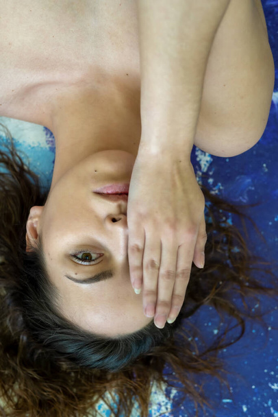 Portrait d'une jeune femme sexy aux cheveux longs brune couchée sur un sol bleu turquoise artistique peint studio, un œil recouvert de sa main
. - Photo, image