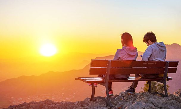 zwei Personen, ein Paar, das zusammen auf einer Bank auf dem Gipfel eines Berges sitzt und den Sonnenuntergang während der goldenen Stunde betrachtet - Foto, Bild