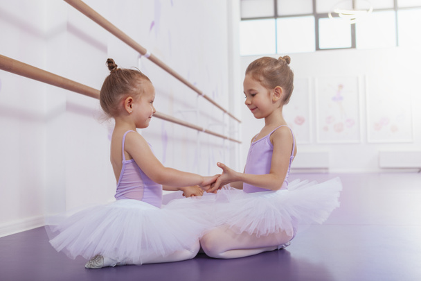 Дві чарівні маленькі балерини тримають руки посміхаючись один одному в балетній школі. Милі щасливі молоді артисти балету відпочивають після тренування
 - Фото, зображення