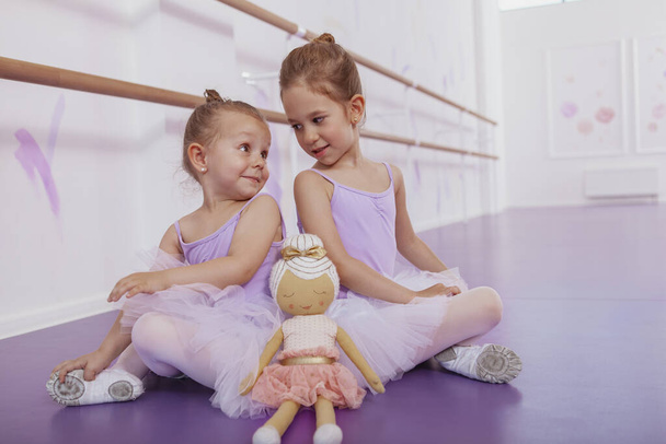 Twee schattige ballerina 's achterover leunend op de grond, elkaar aankijkend over hun schouders. Schattige meisjes op balletles, kopieerruimte - Foto, afbeelding