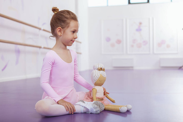Charming young ballerina in pink leotard sitting on the floor with her ballerina doll, resting at ballet school after exercising - Photo, Image
