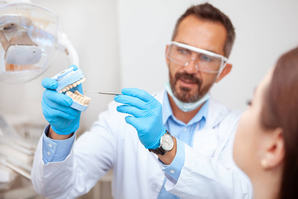 Досвідчений зрілий чоловік-стоматолог пояснює гігієну зубів своєму пацієнтові, тримаючи форму для зубів
 - Фото, зображення