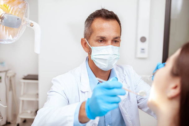Профессиональный стоматолог в медицинской маске, осматривающий зубы пациента
 - Фото, изображение
