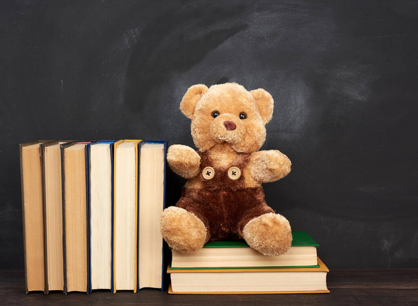 Brauner Teddybär sitzt auf einem braunen Holztisch, hinter einer leeren schwarzen Kreidetafel, zurück zur Schule - Foto, Bild