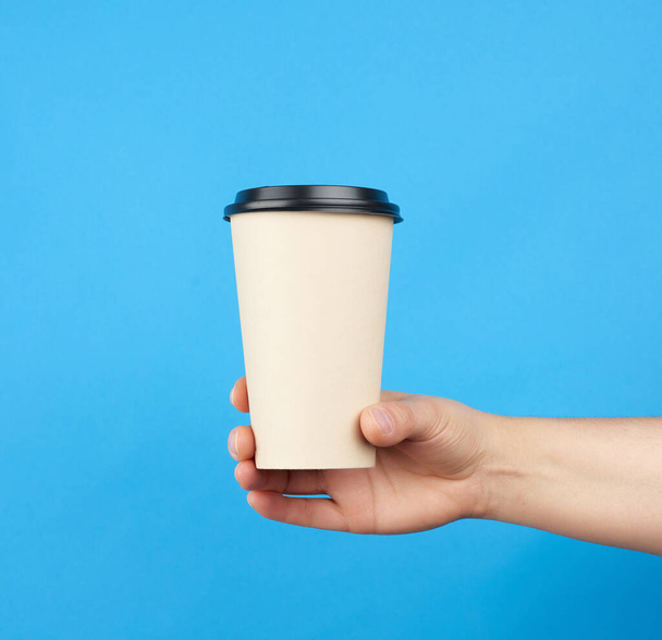 barna papír eldobható csésze műanyag fedéllel, női kéz tartja a kék háttér - Fotó, kép