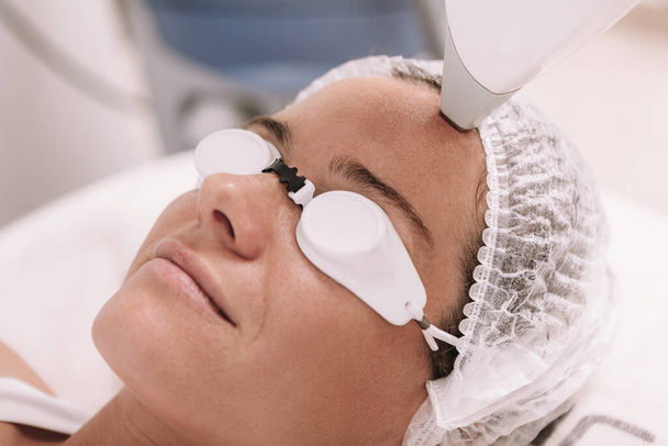 Cropped close up of a mature woman smiling, getting facial hair removed with laser at beauty clinic. Cosmetologue enlever les poils excessifs sur le front d'une cliente
 - Photo, image