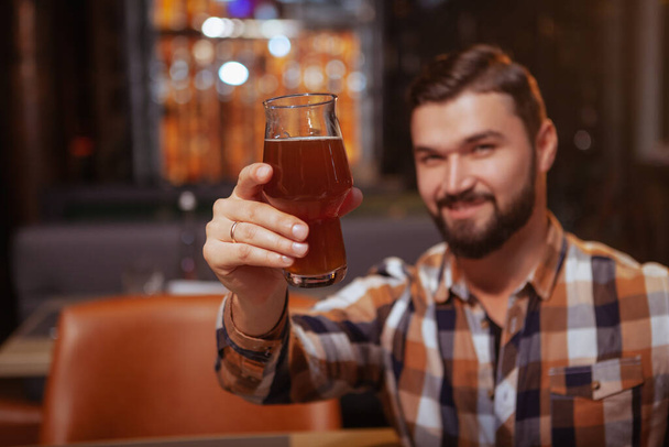 Foco seletivo em um copo de cerveja na mão de homem barbudo bonito feliz. Homem alegre brindando com seu copo para a câmera
 - Foto, Imagem