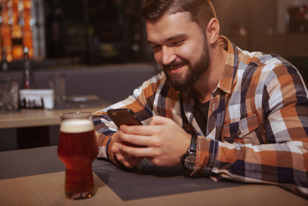 Feliz hombre guapo enviando mensajes en su teléfono inteligente en el pub de cerveza. Atractivo hombre alegre usando su teléfono mientras bebe cerveza en el bar
 - Foto, imagen