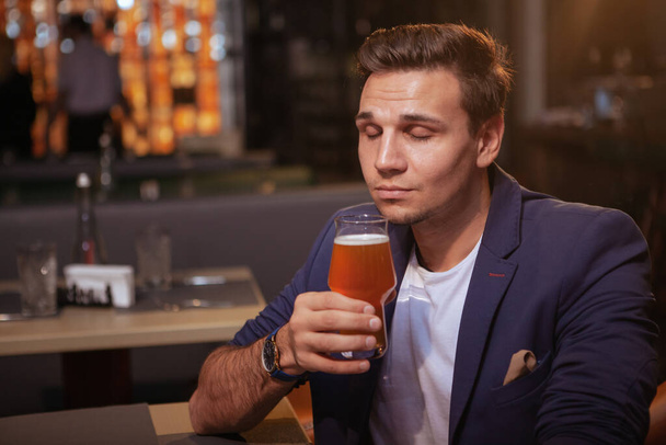 Γοητευτικός νεαρός επιχειρηματίας μυρίζοντας άρωμα νόστιμης μπύρας στο ποτήρι του στο μπαρ - Φωτογραφία, εικόνα