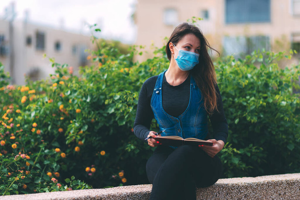 Young woman in a blue medical mask sits in a park with pen in her hand and blank notebook on park. Life after quarantine during the coronavirus epidemic pandemic. Life with a clean slate. New reality - Фото, изображение