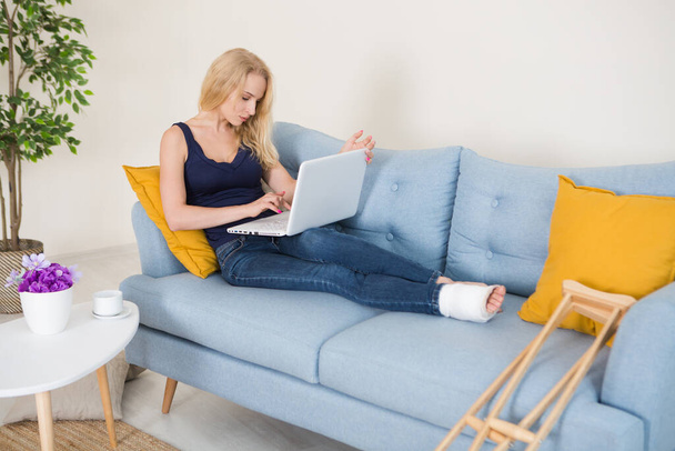Mladá žena se zlomenou nohou v sádře sedí doma na pohovce a pracuje na laptopu. Nemocná žena mluví s doktorem online. konzultace s lékařem online - Fotografie, Obrázek