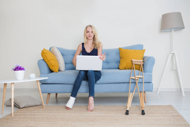 Young woman with  broken leg in cast sitting on sofa at home working on the laptop. Sick woman talks to doctor online. consultation of the doctor online - Foto, Bild