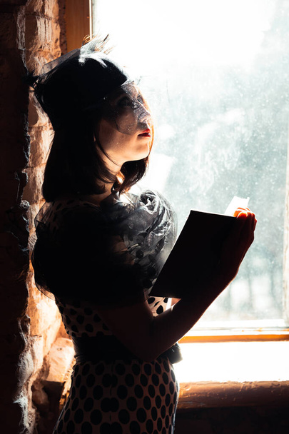 Mädchen im Stil der zwanziger Jahre des letzten Jahrhunderts mit schwarzem Hut, Schleier und Schal auf offenen Schultern am Fenster stehend mit einem Buch in der Hand. - Foto, Bild