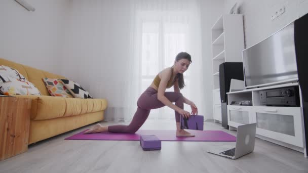 Young beautiful woman is practicing stretch legs on violet mat, she using two bricks to support and looking to the laptop with online instruction - Séquence, vidéo