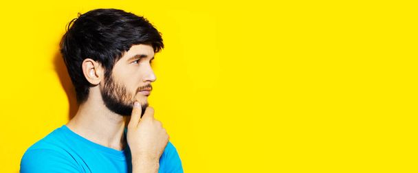 Studio portret van jonge bedachtzame man aanraken kin, op zoek naar lege achtergrond van gele kleur met kopieerruimte. - Foto, afbeelding
