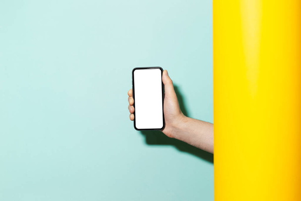 Close-up of male hand holding modern smartphone with white mockup between two studio backgrounds of yellow and aqua menthe colors.  - 写真・画像