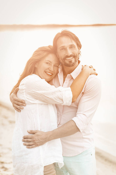 Happy couple hugs on the beach. Smiling man and woman in white clothes looking at camera while standing embracing on sea coast against backdrop of water. Toned image - 写真・画像