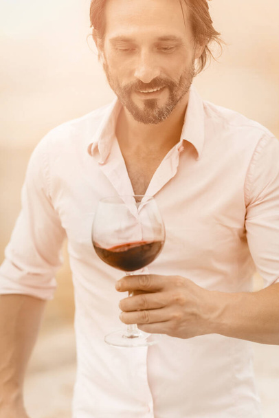 Handsome man going to taste luxury red wine. Winemaker smiles looking for drink in wineglass while standing on the coast. Toned image - Foto, Bild