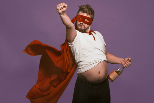 Drunk super antihero holding wine bottle showing his tummy. Man in a red mask and a superhero cloak with bottle of wine in one hand and raising his fist forward. Isolated on grape purple background - Foto, Bild