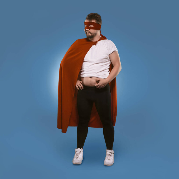 Body positive Super hero man added weight over winter. Sad man in superhero costume checks fat on his tummy. Isolated on blue background. Template with place for text. Overweight concept - Photo, Image