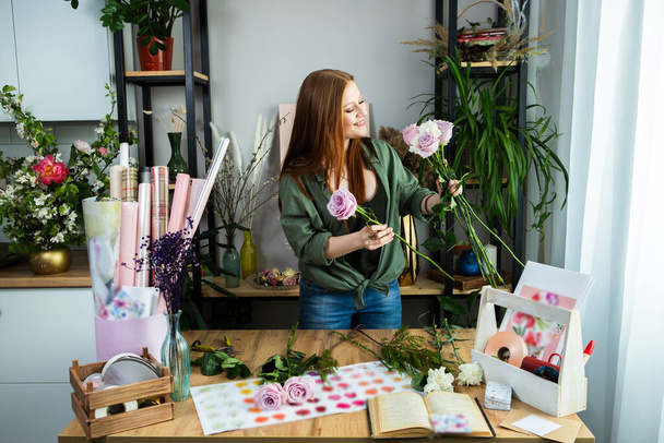 A girl florist with red hair collects a bouquet of roses in a flower shop. Remote workshop for the Assembly of bouquets. - 写真・画像