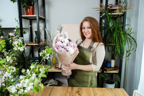 Beautiful florist girl with red hair and a beautiful bouquet of roses at the counter in a flower shop. Sale of flowers and plants - Foto, afbeelding