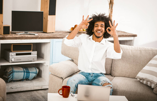 Positive guy shows OK gestures with two hands. Young man toothy laughs at camera while sitting on sofa at coffee table with laptop computer and cup of tea or coffee on it. Toned image - Φωτογραφία, εικόνα