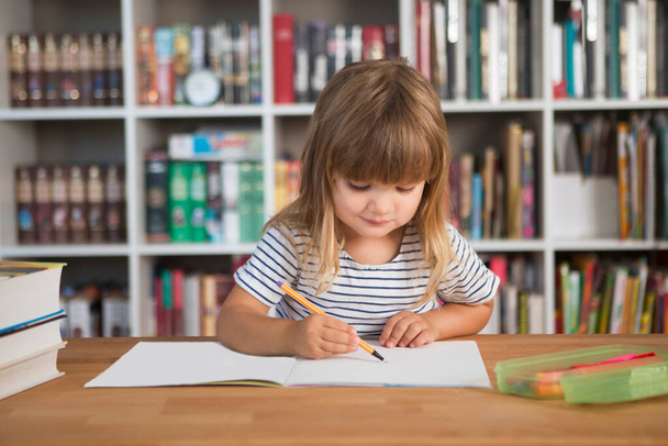 child do lessons at home.  kid study at home.  quarantine. Corona Virus. schoolgirl  study online at home. homeschooling, distant learning. schoolkid drawing at the desk with a bookcase against the background of - Photo, image