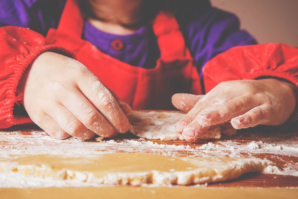 The concept of homemade pastry. Portrait of cute little child girl chef cooking cakes dough on wooden desk with flour.  - Photo, Image