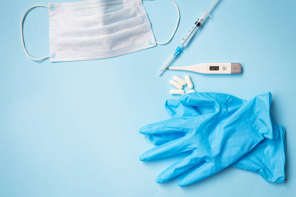 Blue latex gloves on a blue background lie with pills, medical mask, thermometer and syringe - Фото, зображення