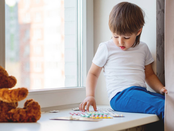 Toddler sits on windowsill and plays with scattering pills without parent's control. Dangerous situation with little boy. Medicines are freely available to child. - Foto, Imagen