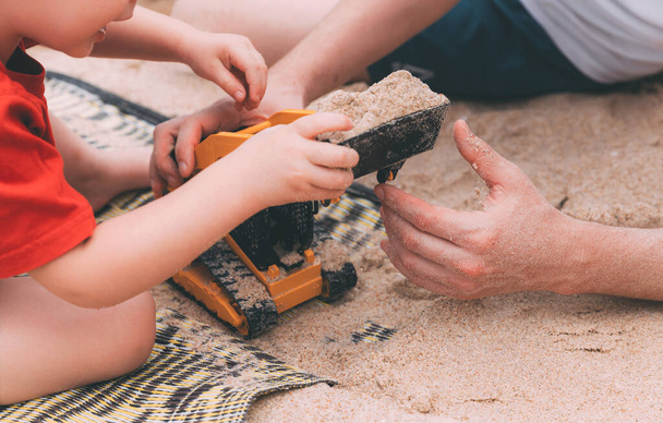 Father's day. Dad and son. Little son plays toys with dad on the beach with sand. Happy loving family. Happy family father and child. Toy cars. Hands of a man and a small child on the beach. Together - Zdjęcie, obraz
