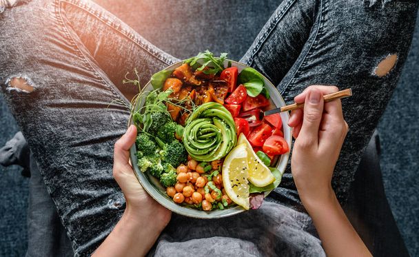 Frau in Jeans hält Buddha-Schüssel mit Salat, gebackenen Süßkartoffeln, Kichererbsen, Brokkoli, Gemüse, Avocado, Sprossen in den Händen. Gesunde vegane Ernährung, saubere Ernährung, Diät, Draufsicht - Foto, Bild
