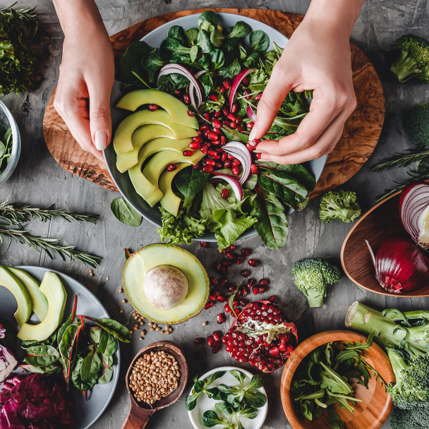 Frau kocht gesunden frischen Salat mit Avocado, Gemüse, Rucola, Spinat im Teller vor grauem Hintergrund. Gesunde vegane Ernährung, saubere Ernährung, Diät, Draufsicht, Straffung - Foto, Bild