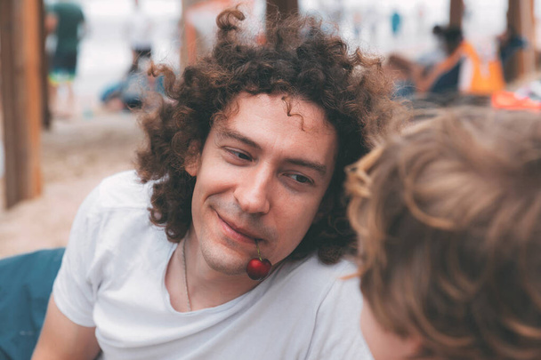 Father's day. Dad and son. Little son feeds dad ripe cherries on the beach. Happy family father and child. Young handsome man with long curly hair smiles and his son in the foreground from back - Фото, изображение
