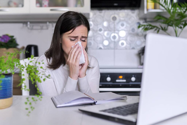 Sick young woman. Sneezing and coughing with handkerchief, sitting with laptop - 写真・画像